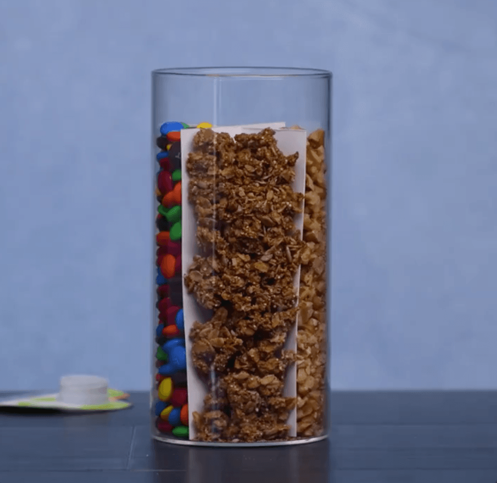 A plastic jar has the cardboard placed inside of it. In each quarter is a different type of snack.